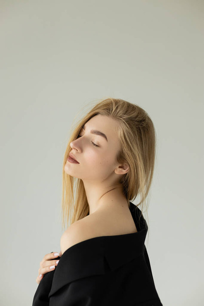 blonde and seductive woman in black blazer posing with closed eyes and bare shoulder isolated on grey - Foto, imagen