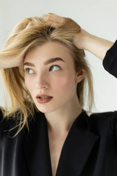 portrait of pretty woman in black jacket adjusting blonde hair and looking away isolated on grey - Foto, immagini