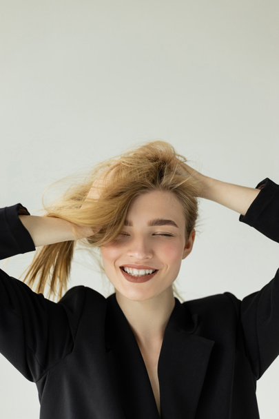 joyful woman in black jacket touching blonde hair and smiling with closed eyes isolated on grey - Photo, Image