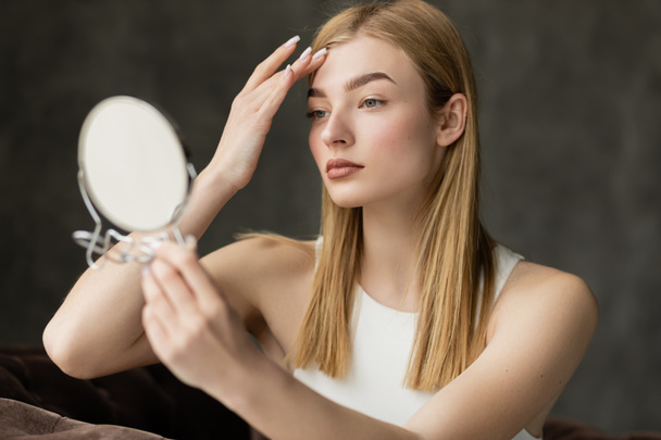 young blonde woman with natural makeup touching forehead while looking in blurred mirror at home - Photo, Image