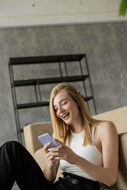 Genç heyecanlı kadın akıllı telefona bakıyor ve oturma odasındaki kanepenin yanında gülüyor. - Fotoğraf, Görsel