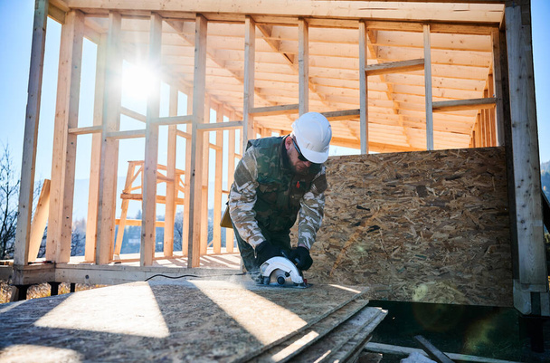 Carpenter using circular saw for cutting wooden OSB board. Man worker building wooden frame house on pile foundation. Carpentry concept. - Fotoğraf, Görsel