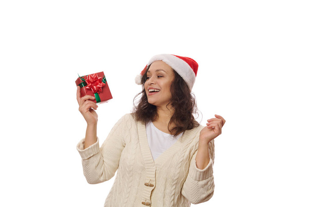 Belle femme afro-américaine portant un chapeau de Père Noël et un pull beige, se réjouissant du cadeau de Noël sur fond blanc. Le jour de la boxe. Il est temps d'ouvrir les coffrets cadeaux. Préparatifs du Nouvel An. Le temps de Noël - Photo, image