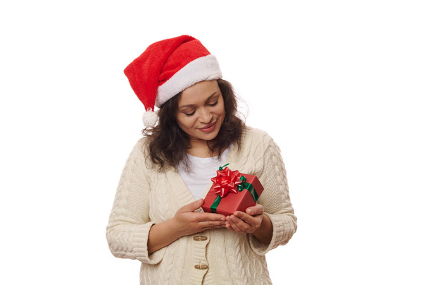 Pleasant woman wearing Santa hat and warm beige sweater, holding Christmas present, wrapped in wrapping gift paper and tied with green shiny ribbon and red bow, isolated on white background. Ad space - Photo, Image