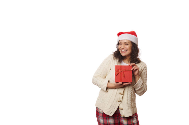 Portret w pasie na białym tle wieloetnicznej ładnej kobiety w kapeluszu Mikołaja, radośnie uśmiechniętej patrząc na przestrzeń reklamową kopii, pozującej z pudełkiem prezentów z prezentem świątecznym prezentem - Zdjęcie, obraz