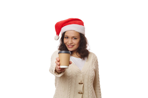 Mujer atractiva multiétnica en el sombrero de Santa, sonriendo misteriosamente y mirando a la cámara mientras sostiene un té caliente para llevar o café de la taza de papel ecológico, aislado en el fondo blanco. Bebida para llevar - Foto, imagen