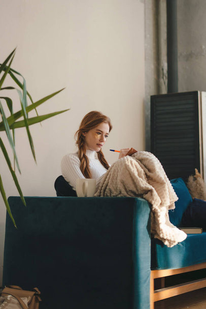 Молодая женщина с карандашом на дневнике сидит на диване на уютной домашней ноте. Перерыв от выгорания и цифровой перегрузки - Фото, изображение