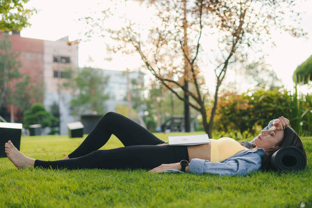 Vista lateral de una chica deportiva descansando y leyendo un libro interesante, al aire libre en el parque de la ciudad. Hermoso verde. Mujer de fitness. Naturaleza verano. Actividades de verano - Foto, imagen