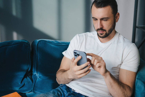 Cheerful handsome young man with under eye patches using his smart phone while sitting on the couch at home. Text message. Happy lifestyle. - Φωτογραφία, εικόνα