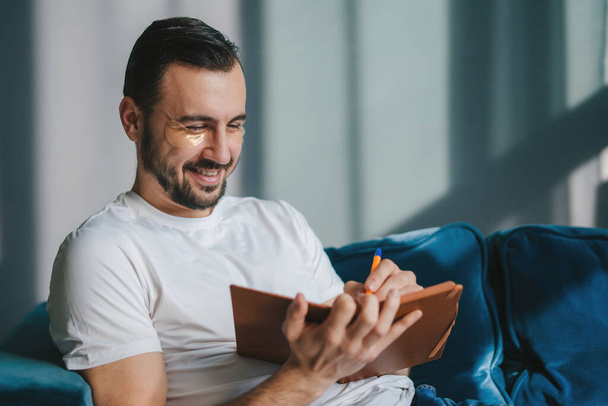 Hymyilevä nuori opiskelija mies valkoisessa t-paidassa istuu sinisellä sohvalla sisätiloissa kirjoittamassa muistiinpanoja muistikirjaan, lepäämässä viikonloppuisin kotona. Onnellinen - Valokuva, kuva