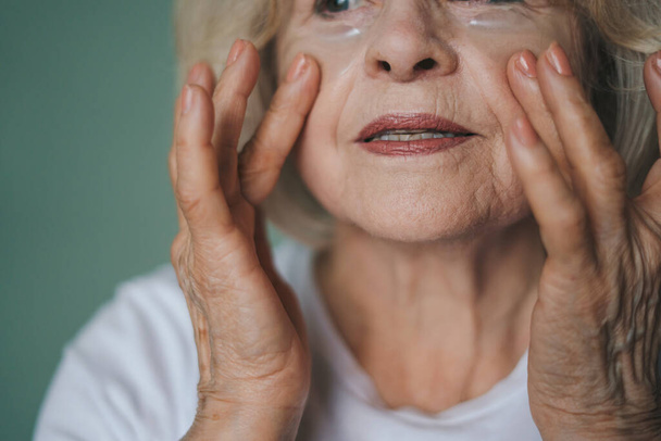 Lähikuva muotokuva eläkeläinen nainen soveltamalla ravintoa silmien alla laastaria etsivät tuoretta aamulla. Kauneus, ihonhoito ja vanhusten käsite - Valokuva, kuva