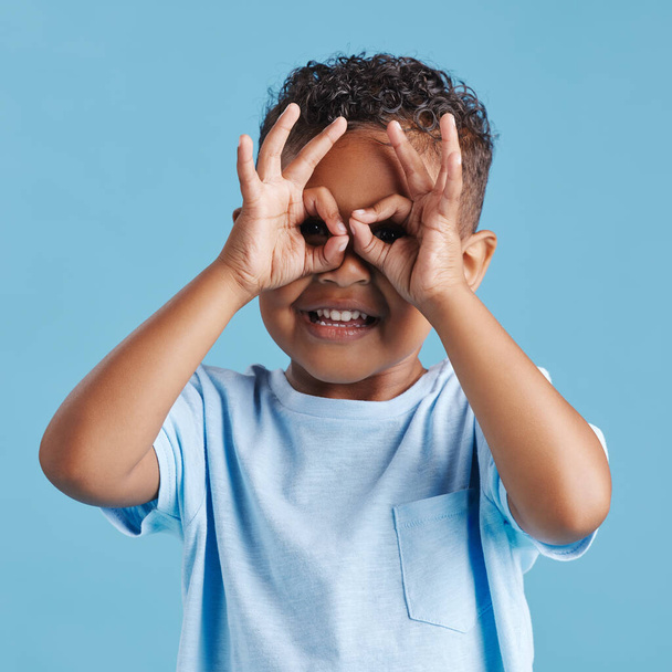 Портрет любознательного любопытного маленького мальчика, смотрящего сквозь пальцы в форме бинокля на фоне синей студии. Любопытный ребенок исследует. - Фото, изображение