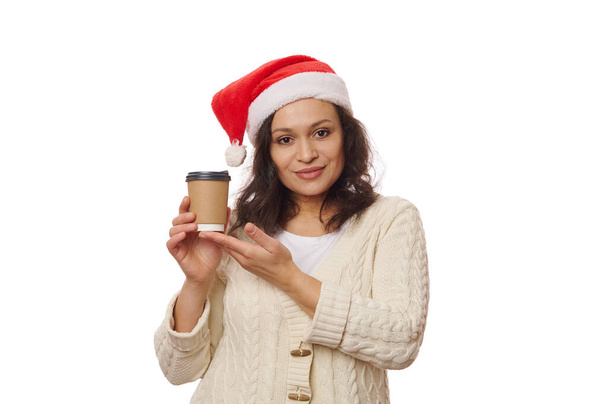 白い背景に隔離され、魅力的な多民族のブルネットの女性は、サンタの帽子をかぶって、リサイクル可能な使い捨てエコペーパーカップで持ち帰りの熱いお茶やコーヒーを持っています。飲食消費主義. - 写真・画像
