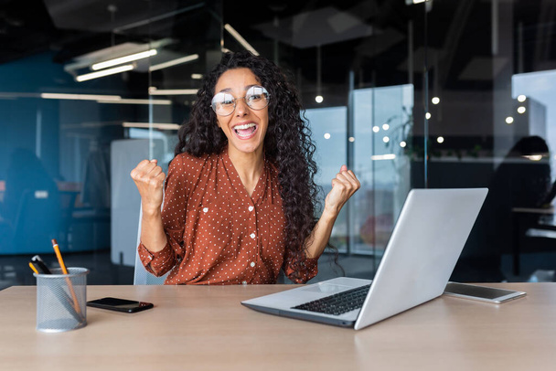 Portrait successful Hispanic business woman office worker with curly hair smiling and looking at camera working at table with laptop holding hand up gesture success and triumph celebrating victory - Foto, imagen