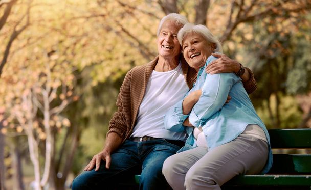 Senior couple, park bench and happy while sitting together in retirement for freedom, peace and calm with a smile and happiness in nature. Old man and woman outdoor to relax while on vacation. - Foto, Imagen