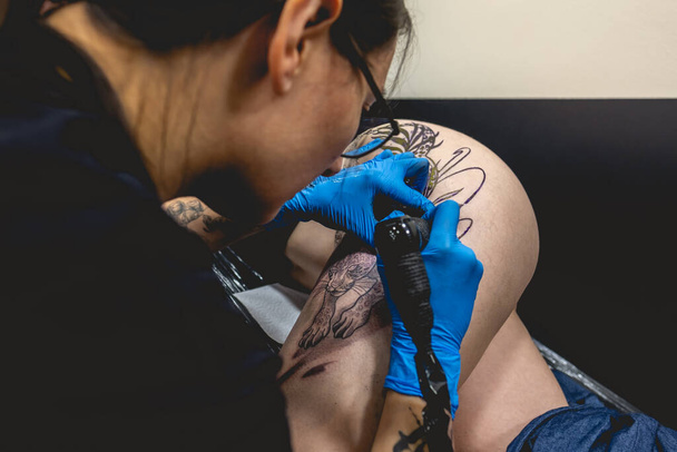 Dövmeli genç bir kız dövme makinesiyle dövme yapıyor. Büyük bir kaplanın kalçasında ve güzel bir kadının kalçasında. - Fotoğraf, Görsel