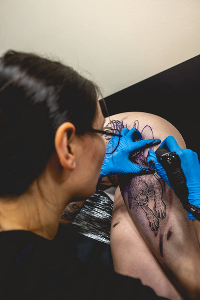 Νεαρή καλλιτέχνης τατουάζ κορίτσι με γυαλιά κάνει ένα τατουάζ, με μηχανή τατουάζ, μιας μεγάλης τίγρης, στο μηρό και τους γοφούς μιας όμορφης γυναίκας - Φωτογραφία, εικόνα