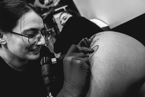 Mosolygó fiatal tetováló művész lány szemüveges, hogy egy tetováló gép, egy nagy tigris, a comb és a csípő egy gyönyörű nő (fekete-fehér) - Fotó, kép