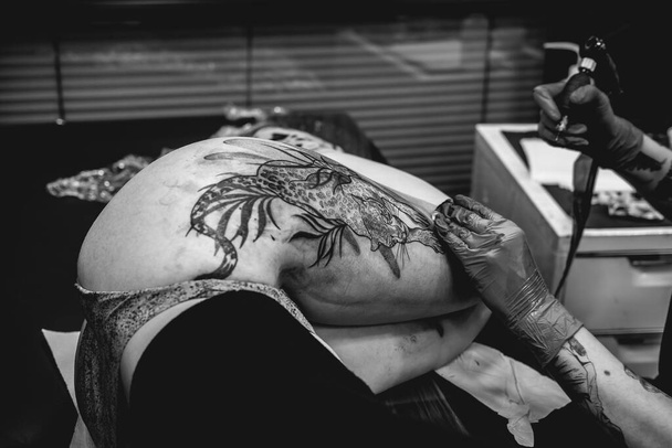 Tattoo artist with blue gloves making a tattoo, with tattoo machine, of a big tiger, in the thigh and hips of a beautiful woman enduring pain (in black and white) - Photo, Image