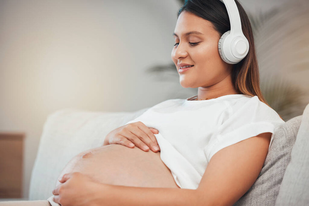 Nő, terhes és pihenjen, miközben hallgat nyugodt zenét kanapén egészséges zen, wellness vagy meditáció otthon. Terhes nő mosoly relaxációs tartja has fülhallgató a nappaliban kanapé. - Fotó, kép
