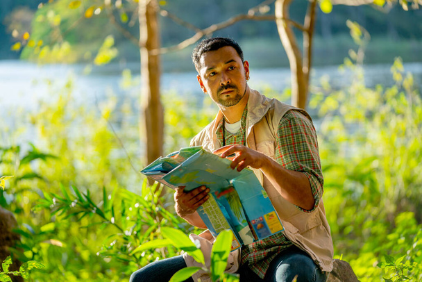 Азиатский мужчина держит карту и сидеть в лесу с расслабиться во время путешествия и кемпинга в национальном парке. - Фото, изображение