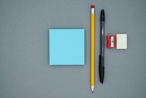 Bleistift, Stift, Schaber, Radiergummi und blaue Blanko-Notizen auf grauem Hintergrund, Draufsicht. Schulbürobedarf. - Foto, Bild