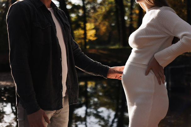 cropped photo of couple, man and pregnant wife are holding hands and walking by the lake on sunlight sunset. Happy family enjoying pregnancy on nature. The concept of Mother's Day and Women's Day - Photo, Image