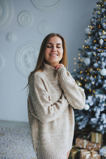 Egy fiatal, szőke hajú nő, mosollyal az arcán, kötött pulcsiban áll egy díszített karácsonyfa mellett. Az új év ünneplése. - Fotó, kép