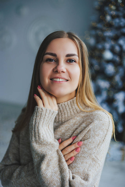 Молодая светловолосая женщина с улыбкой на лице в трикотажном свитере стоит рядом с украшенной елкой. Празднование нового года. - Фото, изображение
