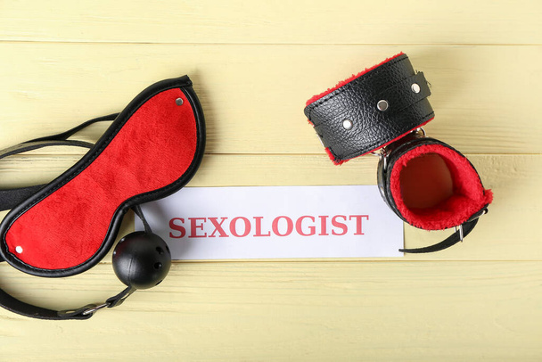 Wort SEXOLOGIST mit Handschellen, Mundknebel und Schlafmaske auf gelbem Holzgrund - Foto, Bild
