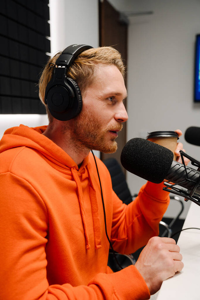 Happy young male radio host using microphone and headphones while broadcasting in studio - Foto, afbeelding