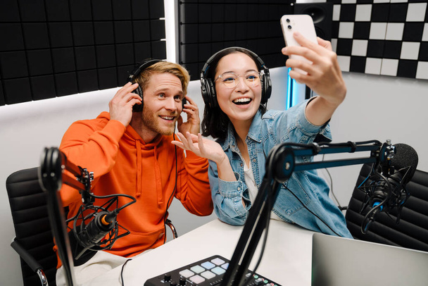 Happy young man and woman smiling and taking selfie photo while broadcasting in radio studio - Foto, afbeelding