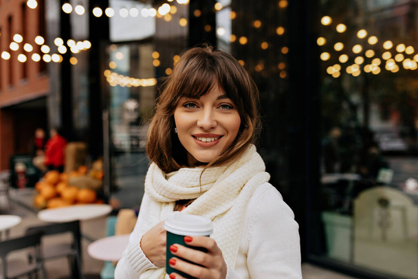 Adorable smiling happy european girl with wavy hair and wonderful smile drinking coffee outside on background of city cafe. She wears white sweater and scarf. - Photo, Image