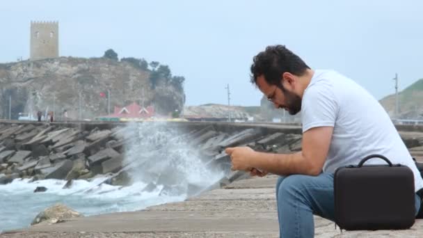 człowiek siedzący na wybrzeżu, troskliwy mężczyzna siedzący na wybrzeżu i korzystający z telefonu - Materiał filmowy, wideo