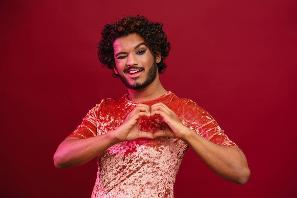 Young curly man with makeup winking while making heart gesture isolated over pink background - Photo, image