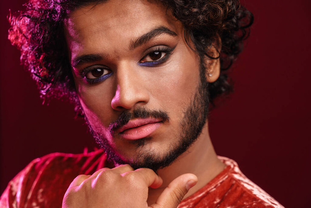 Young curly man with makeup posing and looking at camera isolated over red background - Foto, Imagen