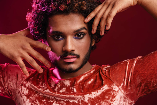 Young curly man with makeup gesturing and looking at camera isolated over red background - Фото, зображення