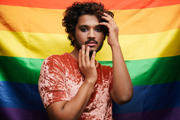Young curly man with makeup posing and looking at camera isolated over rainbow background - Fotó, kép