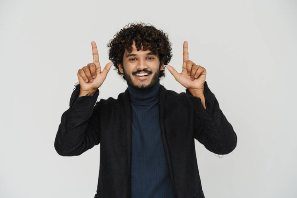 Adult young indian man smiling and pointing with fingers and raised arms looking at camera over isolated grey background - Valokuva, kuva