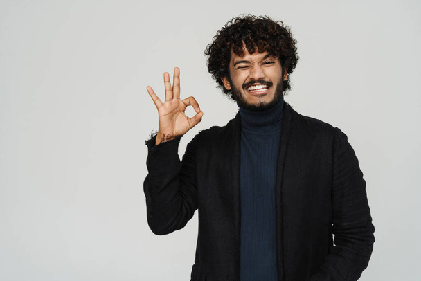 Young handsome curly smiling and winking indian man showing ok gesture over isolated grey background - Valokuva, kuva