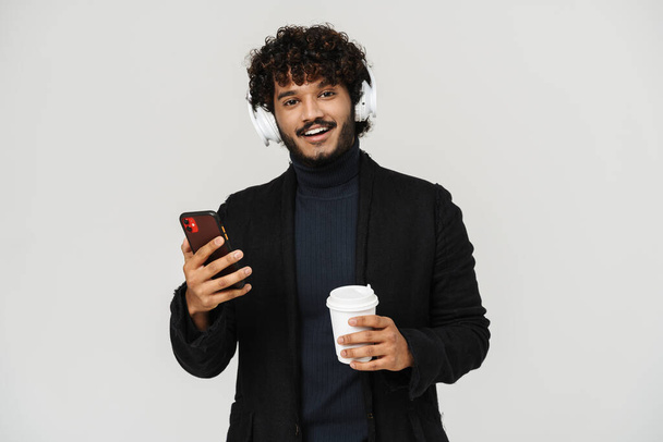 Young adult indian smiling curly man in headphones with coffee and phone looking on camera over igrey isolated background - Fotografie, Obrázek
