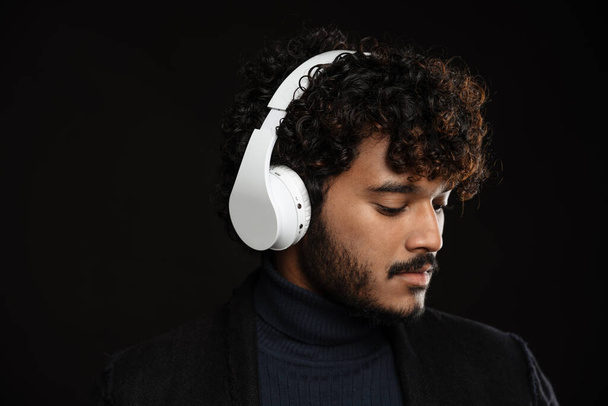 Portrait of young indian curly pensive man in headphones looking downwards over isolated dark background - Φωτογραφία, εικόνα