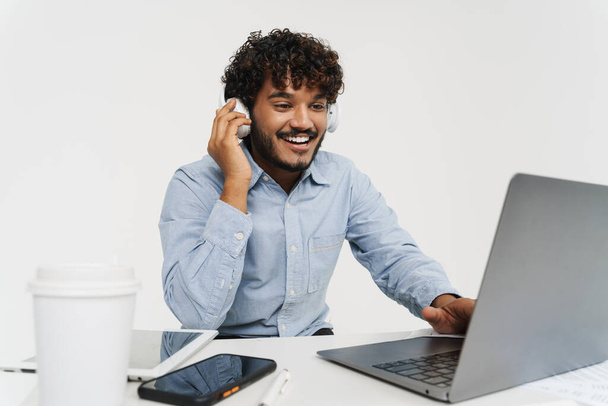 Adult indian curly smiling office employee sitting with laptop in headphones over isolated grey background - Foto, Imagem
