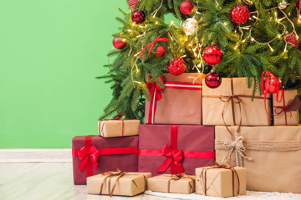 Cadeaus onder gloeiende kerstboom in de buurt van groene muur - Foto, afbeelding