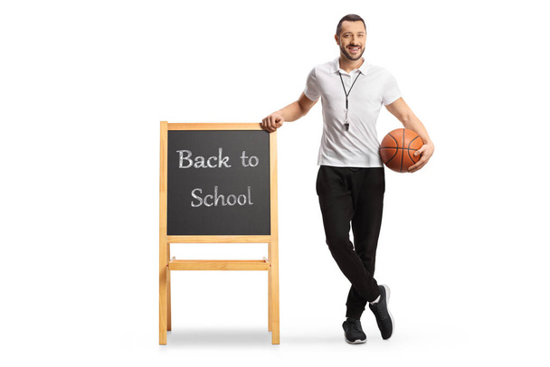 Physical education teacher holding a basketball and leaning on a blackboard with text back to school isolated on white background - Photo, image