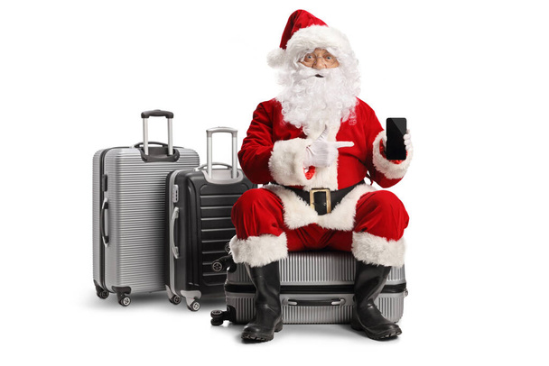 Excited santa claus sitting on a suitcase and pointing at a smartphone isolated on white background - Zdjęcie, obraz