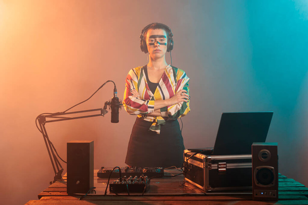 Female performer posing with arms crossed at turntables, using headphones and mixer to produce techno music or sounds. Doing performance with mixer and control buttons, audio stereo equipment. - Photo, Image