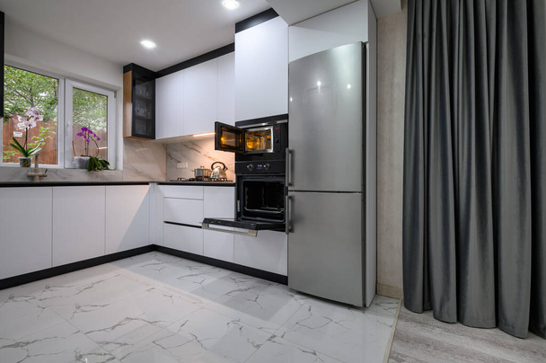 Moderní a funkční kuchyň s bílým designem, mramorovou podlahou, šedou oponou a otevřenými dveřmi do trouby. Ideální pro vaření a těší život. - Fotografie, Obrázek