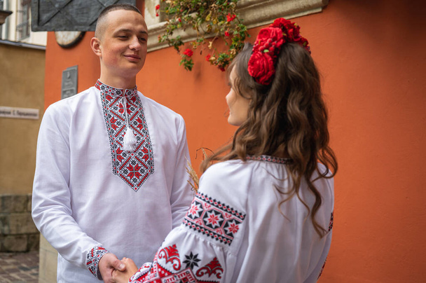 Una feliz pareja joven enamorada, una familia caminando por la vieja ciudad de Lviv en camisas bordadas ucranianas, cogidas de la mano sobre un fondo rojo-naranja. Los jóvenes se abrazan en el casco antiguo de Lviv  - Foto, imagen