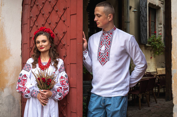 Una giovane coppia felice innamorata, una famiglia che cammina attraverso la città vecchia di Leopoli in camicie ricamate ucraine, tenendosi per mano su uno sfondo rosso-arancio. I giovani si abbracciano nel centro storico di Leopoli  - Foto, immagini